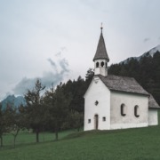 Kościół z białą, poszarzałą elewacją na tle lasu