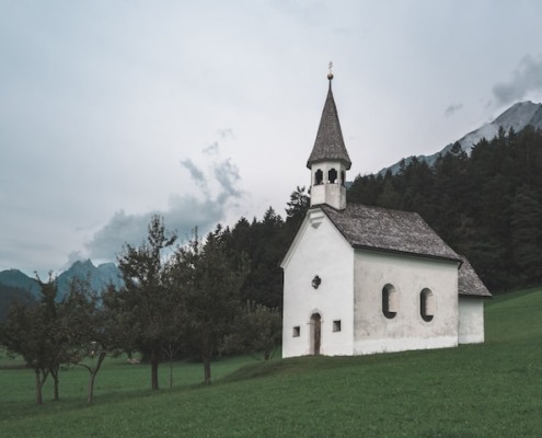 Kościół z białą, poszarzałą elewacją na tle lasu